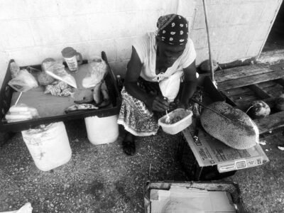 Dar, market lady, has taken a break during a lull at the Saturday Market and is eating lunch on Saturday 29th, 2025 Market lady eating in black and white