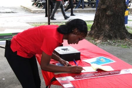 Members writing receipts at the Productions and Operations Management Seminar from the School of Business at the Utech Papine Campus on November 27th, 2025 Girl in red shirt and black pants bending to write receipts on the table
